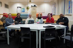 A group of six people participating in the Homebrew Website Club Nuremberg 04/25 seated at a white table in a modern workspace, surrounded by colorful seating, laptops, and a projector mounted on a wooden stand.